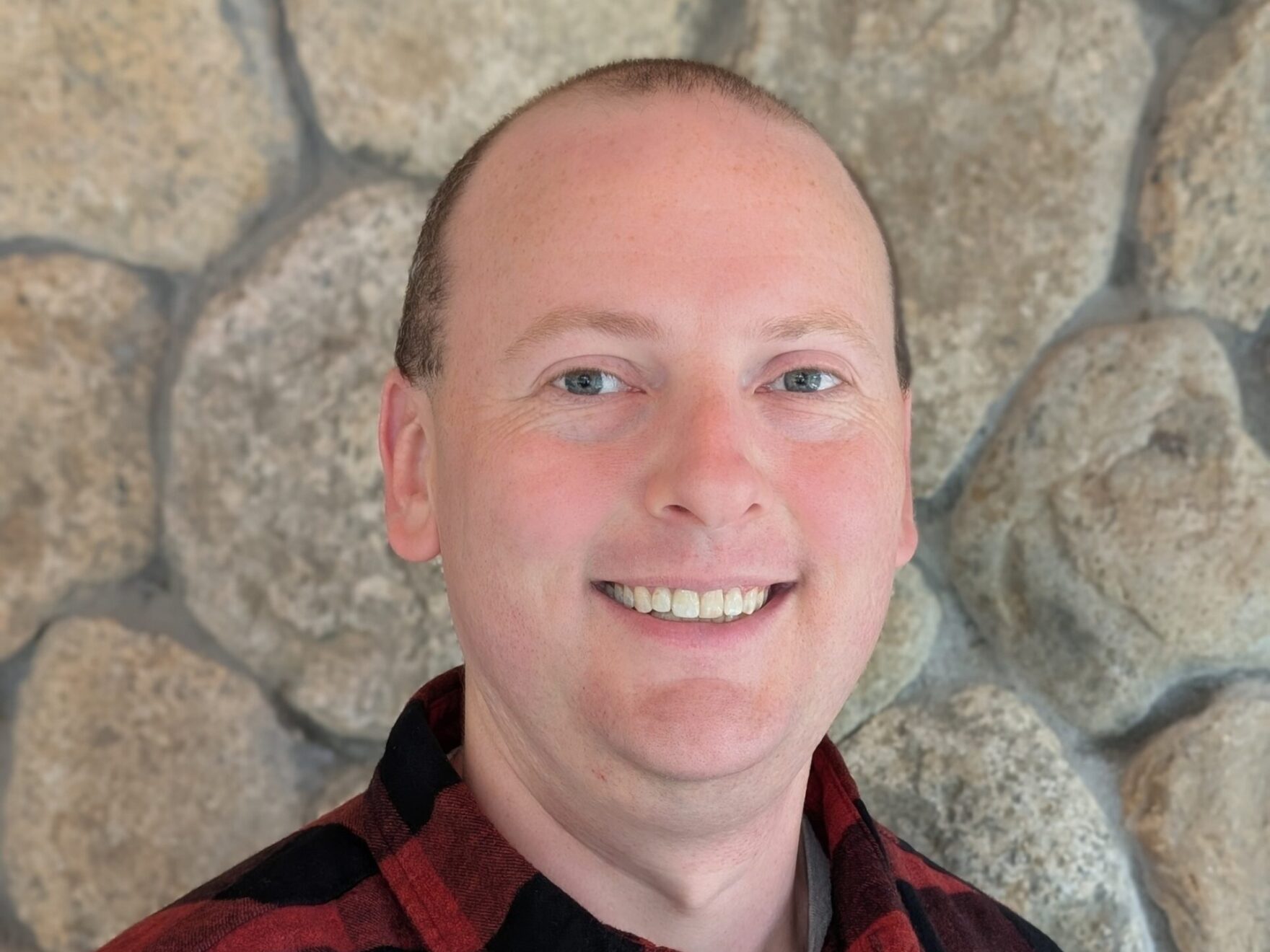 A man stood against a stone walk background wearing a red check shirt and smiling.