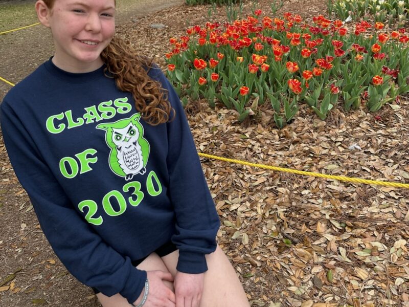 A young teenager sits on the ground in front of flowers smiling at the camera.