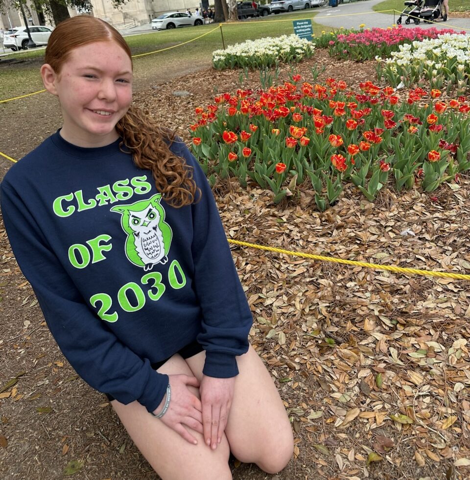 A young teenager sits on the ground in front of flowers smiling at the camera.