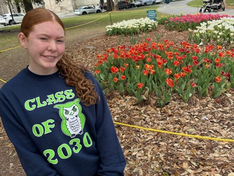 A young teenager sits on the ground in front of flowers smiling at the camera.