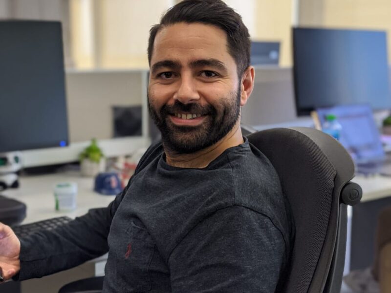 A man with dark hair and a beard faces the camera and smiles in an office environment.
