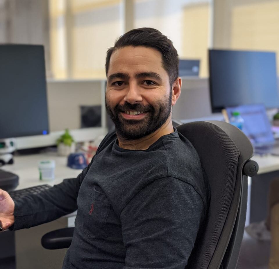A man with dark hair and a beard faces the camera and smiles in an office environment.