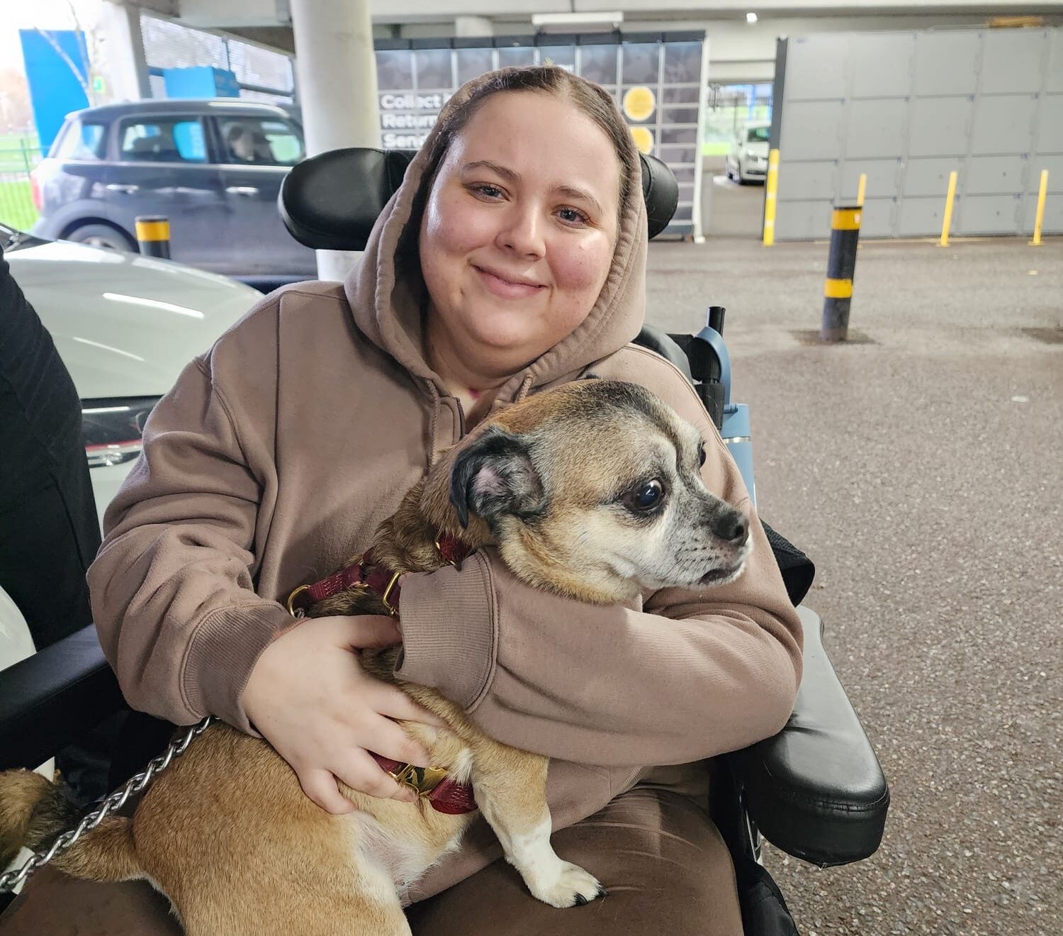 A young woman sits in a wheelchair with a dog on her lap as she smiles to the camera.