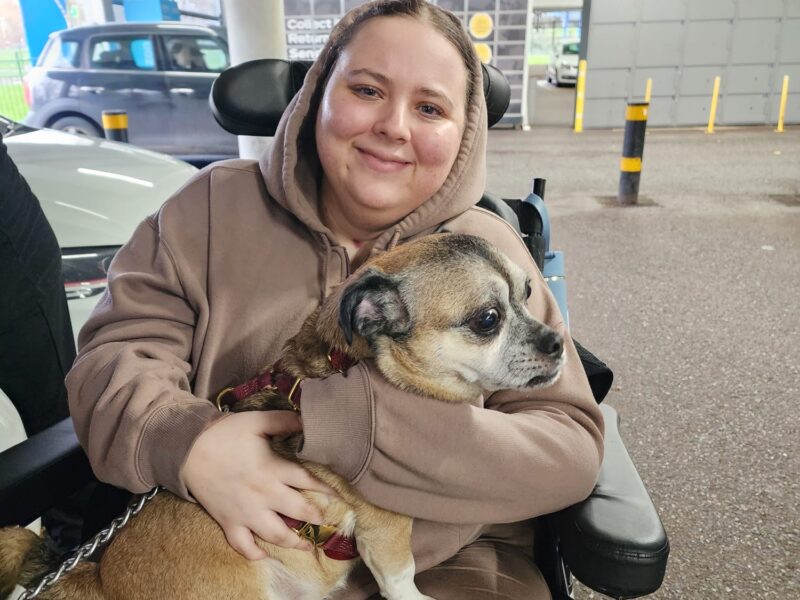 A young woman sits in a wheelchair with a dog on her lap as she smiles to the camera.