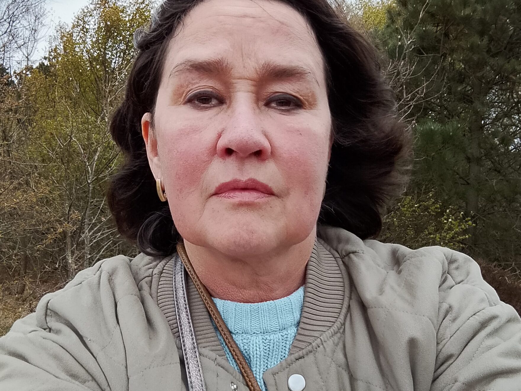 A woman with dark hair and a beige coat looks into the camera.