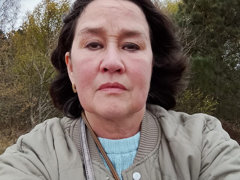 A woman with dark hair and a beige coat looks into the camera.