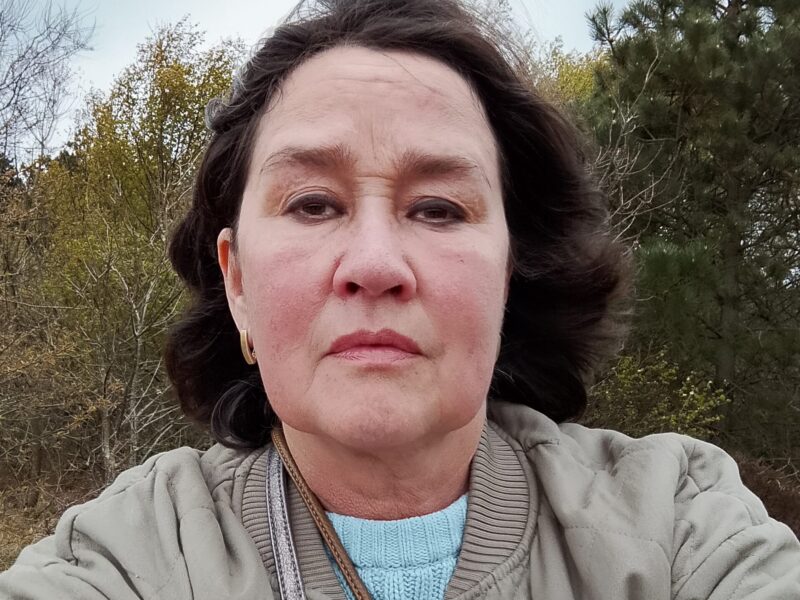 A woman with dark hair and a beige coat looks into the camera.