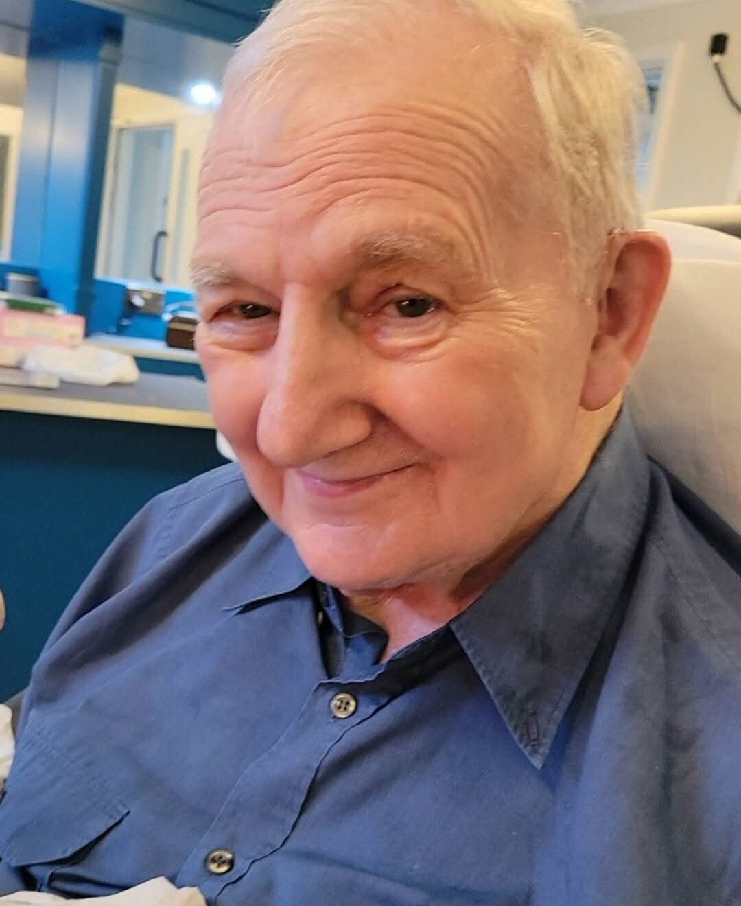 An elderly gentleman wears a blue shirt and smiles at the camera.