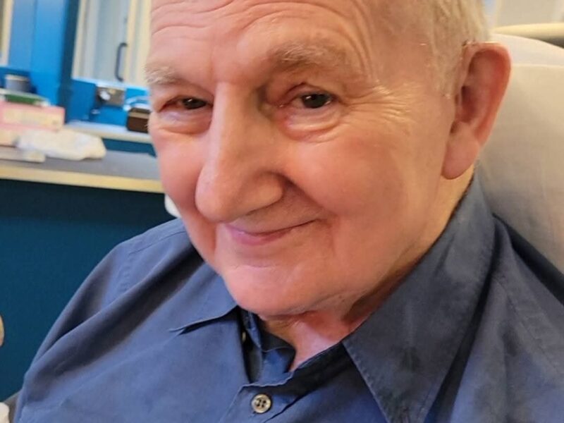 An elderly gentleman wears a blue shirt and smiles at the camera.