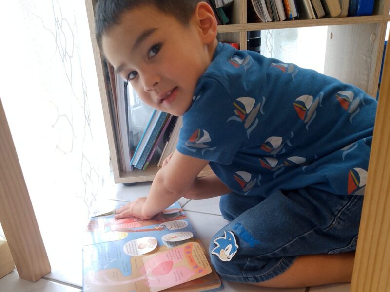 A young boy is reading a book on the floor. He smiles at the camera.