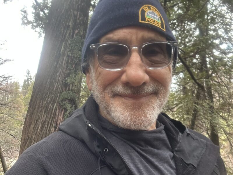 A man with a grey beard wearing a wooly hat and sunglasses smiles at the camera.